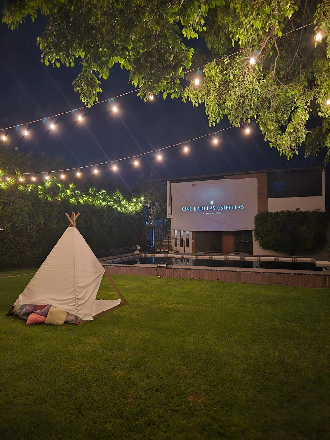 Jardín de noche con teepee, luces Edison colgantes y la pantalla de Cine Bajo las Estrellas junto a la alberca