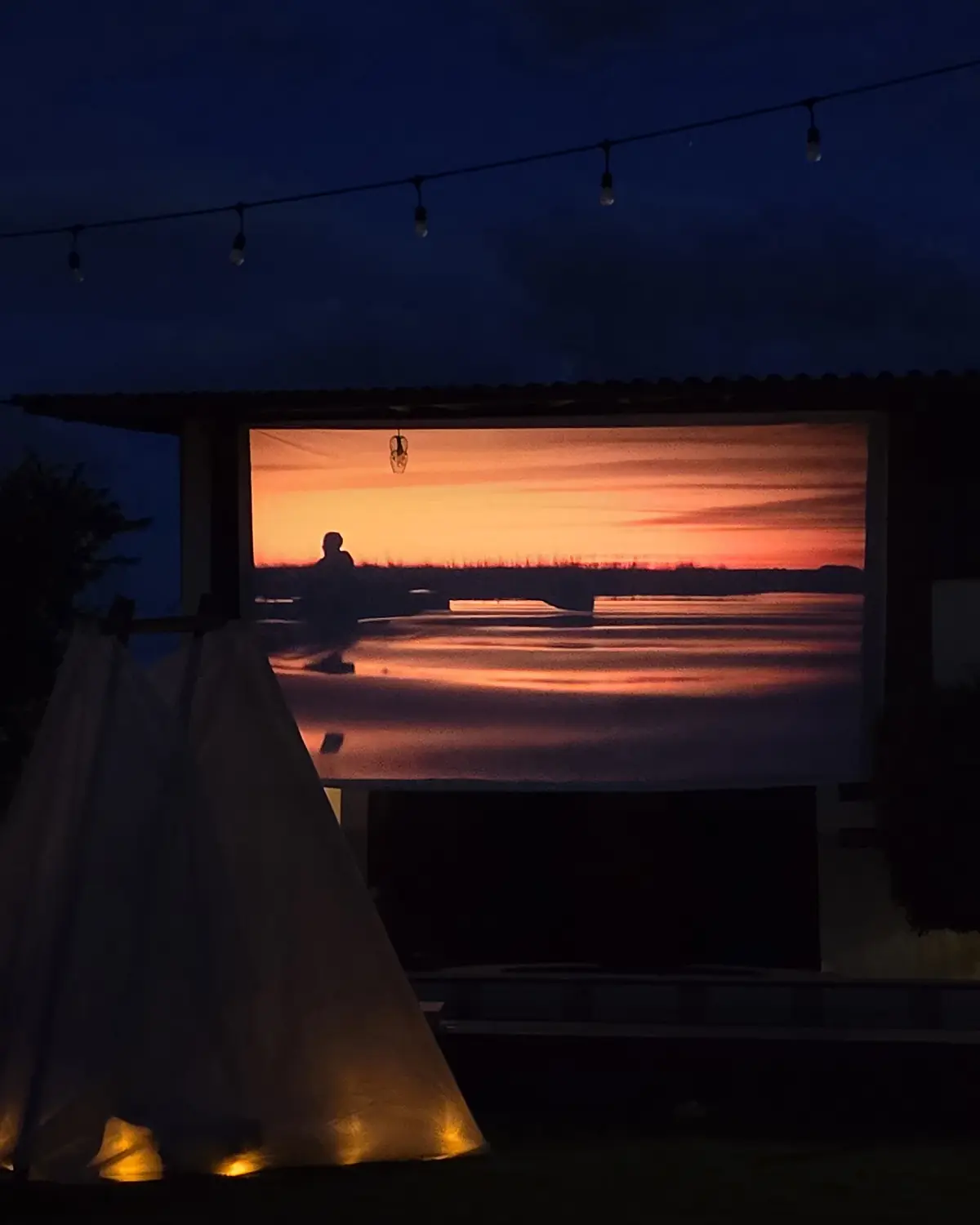El teepee iluminado por dentro frente a la pantalla, con un atardecer proyectado
