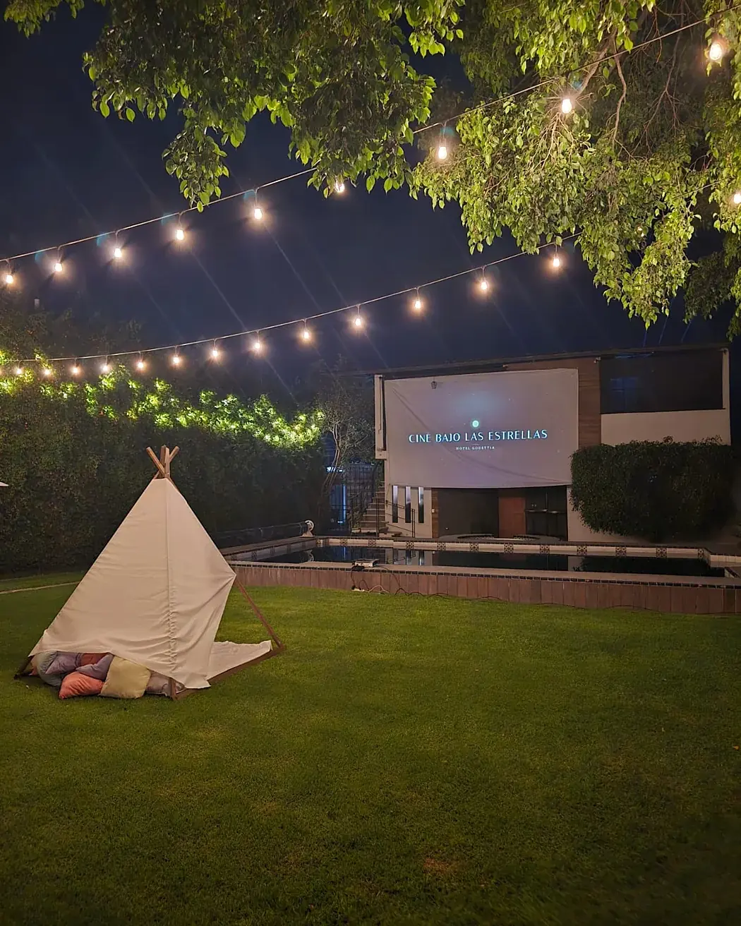 Luces Edison cruzando el jardín bajo el árbol, con el teepee y la pantalla al fondo