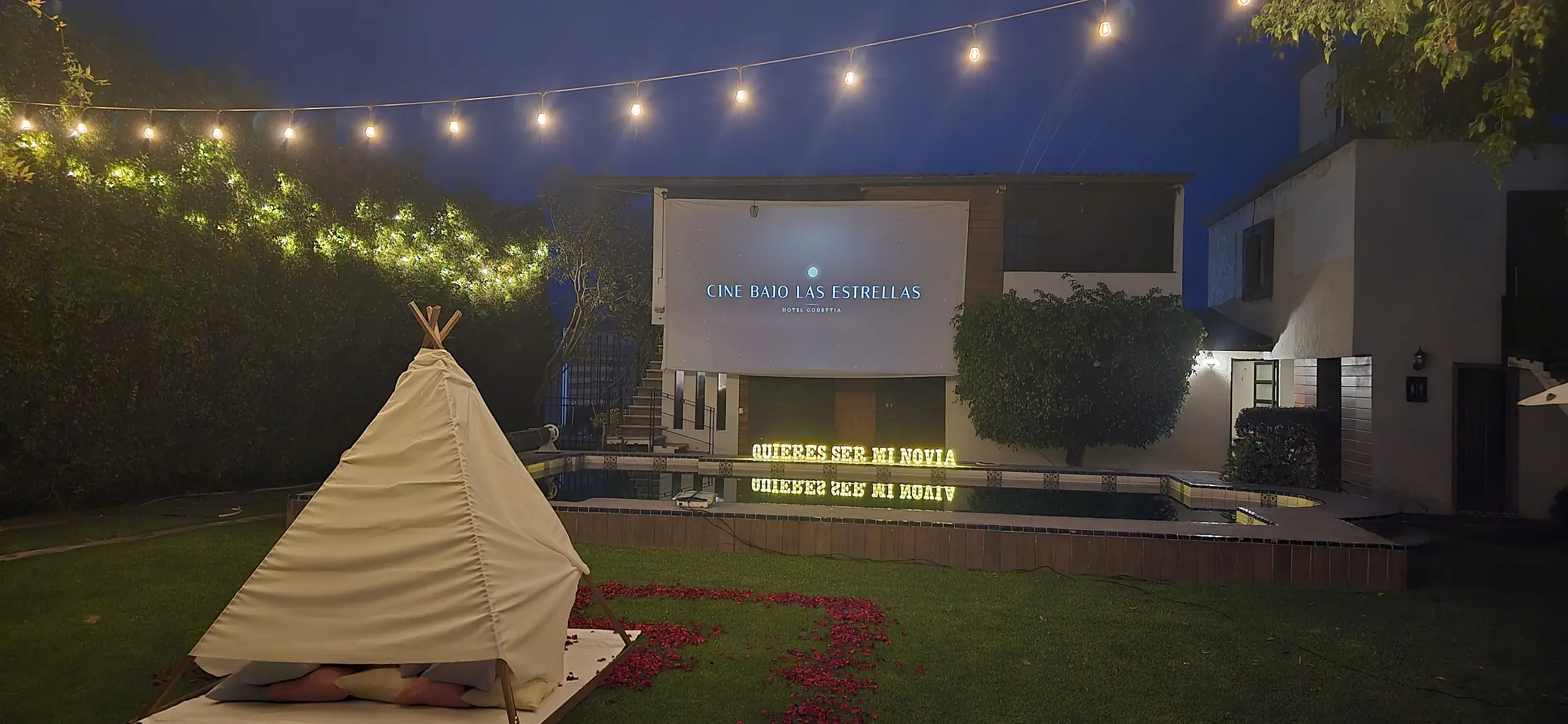 El jardín de noche con el teepee sobre pétalos, las luces encendidas y la pantalla que dice Cine bajo las estrellas